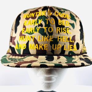 May include: A camouflage trucker hat with the text "HUNTER'S CODE, EARLY TO BED, EARLY TO RISE, HUNT LIKE HELL, AND MAKE UP LIES" embroidered in yellow. The hat features a brown brim and mesh backing.