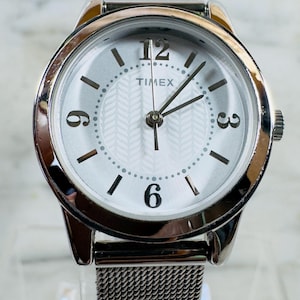 Pode incluir: Relógio Timex prateado com mostrador branco e pulseira de malha. O mostrador apresenta algarismos pretos às 12, 3, 6 e 9 horas, com pequenos pontos marcando as outras horas. Os ponteiros são pretos.