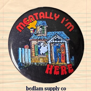 May include: A circular pinback button with a black background featuring a haunted house illustration, a white ghost, and a skeleton in a window. The text reads MENTALLY I'M HERE in red, with the text bedlam supply co printed at the bottom.