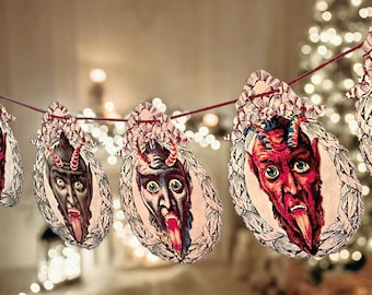 May include: A Christmas garland featuring a string of red and black devil faces with horns and tongues sticking out. The garland is made of paper and is strung on a red string. The garland is hanging in front of a Christmas tree with white lights.