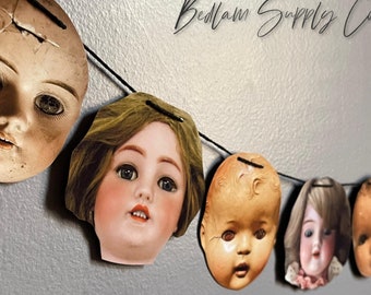 May include: A garland of six porcelain doll heads, each with painted features and hair, strung together with black thread. The garland is hanging against a gray wall. The text "Bedlam Supply Co." is written in black cursive above the garland.
