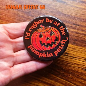 K&ouml;nnte beinhalten: Ein schwarzer Knopf mit einem wei&szlig;en und orangefarbenen K&uuml;rbisdesign. Der Text um den K&uuml;rbis lautet "I'd rather be at the pumpkin patch".