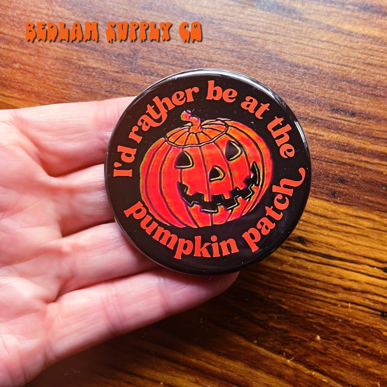 K&ouml;nnte beinhalten: Ein schwarzer Knopf mit einem wei&szlig;en und orangefarbenen K&uuml;rbisdesign. Der Text um den K&uuml;rbis lautet "I'd rather be at the pumpkin patch".