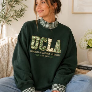 May include: A dark green long-sleeve sweatshirt featuring a mock neck collar and cuffs with a green and white gingham pattern. The front displays the text UCLA in patterned block letters, with University of California, Los Angeles and Est. 1919.
