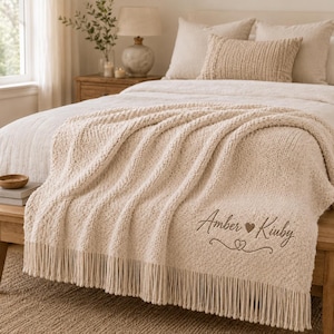 May include: A cream-coloured knitted throw blanket with long fringe trim along the bottom edge. The blanket features embroidered text that reads Amber Kirby with a heart symbol between the names and a decorative heart flourish underneath.
