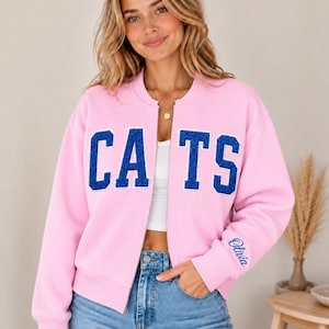 Puede incluir: Una chaqueta bomber rosa claro con cremallera que presenta la palabra CATS en letras grandes de estilo universitario con purpurina azul en el pecho. El puño de la manga izquierda muestra el nombre Olivia bordado en azul. Cuello acanalado.