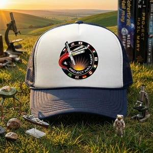 May include: A navy and white trucker hat with a circular patch displaying the text "PROJECT HAIL MARY". The patch features a rocket, flags, and the words "HAIL MARY". The hat has a mesh back and a curved peak.