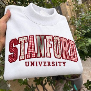 May include: A white crewneck sweatshirt featuring the word STANFORD in large, arched, red-bordered letters. Each letter is filled with a different pattern, including red and white polka dots, gingham, stripes, and floral prints, with UNIVERSITY below.