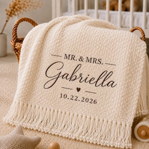 May include: A cream-coloured textured knit throw blanket featuring dark brown embroidered text that reads MR. & MRS. Gabriella, a small heart icon, and the date 10.22.2026. The blanket has a decorative fringe along the bottom edge.