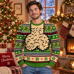 May include: A green and beige long-sleeve knit sweater featuring a repeating holiday pattern of red hearts, white snowflakes, and candy canes, with a large, central graphic of two beige, textured, rounded shapes outlined in black.