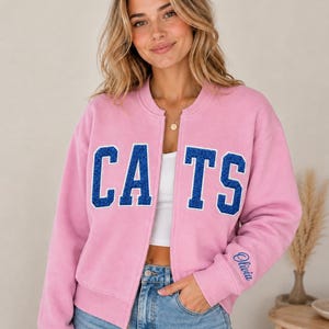 Puede incluir: Una chaqueta bomber rosa claro con cremallera que presenta la palabra CATS en letras grandes de estilo universitario con purpurina azul en el pecho. El puño de la manga izquierda muestra el nombre Olivia bordado en letra azul. Se lleva sobre una prenda blanca.