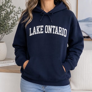 May include: A navy blue hooded sweatshirt featuring the text LAKE ONTARIO printed in white capital letters across the chest. The garment includes a front kangaroo pocket, a drawstring hood with matching navy blue cords, and long sleeves.