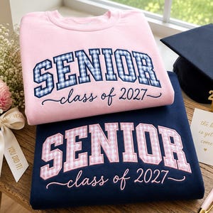 May include: A light pink sweatshirt with blue gingham embroidered text reading SENIOR class of 2027 and a navy blue sweatshirt with pink gingham embroidered text reading SENIOR class of 2027, accompanied by a navy graduation cap with a gold tassel.