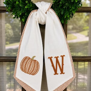 May include: A white fabric door hanger featuring a brown embroidered pumpkin on the left side and a large brown embroidered letter W on the right side. The fabric is finished with a brown and white checkered border along the edges.