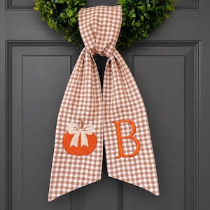 May include: A brown and white gingham fabric door hanger featuring an embroidered orange pumpkin with a cream-colored bow on the left side and a large orange letter B on the right side, suspended from a green leafy wreath on a dark gray door.