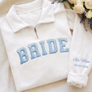 May include: A white quarter-zip sweatshirt featuring the word BRIDE embroidered in light blue block letters across the chest. The sleeve is embroidered with the text Mrs. Wilson and the date 08.06.2026 in a matching light blue script font.