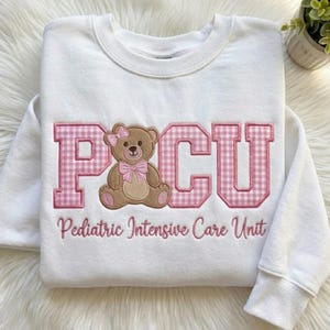 May include: A white long-sleeve sweatshirt featuring the letters PCU in a pink and white gingham pattern. A brown teddy bear with a pink bow on its head and a pink bow around its neck sits in place of the letter C. The text Pediatric Intensive Care Unit is embroidered in pink below the graphic.