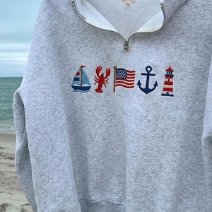 May include: A light gray heathered quarter-zip sweatshirt featuring embroidered nautical icons across the chest, including a sailboat, a red lobster, an American flag, a navy blue anchor, and a red and white striped lighthouse.