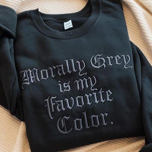 May include: Black sweatshirt with the embroidered text "Morally Grey is my Favorite Color." The long-sleeved top is folded on a beige blanket. The text is in a gothic font.