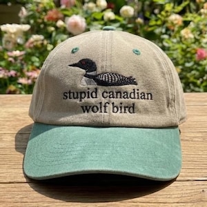 May include: A tan baseball cap with a green bill and top button. The cap features an embroidered loon design and the words "stupid canadian wolf bird". The hat is sitting on a wooden surface with a blurred background of flowers and greenery.
