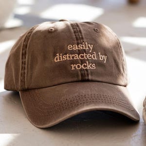 May include: A brown baseball cap with the embroidered text "easily distracted by rocks" in a lighter brown color. The cap has a curved brim and a classic six-panel design. The hat is made of a soft, worn material.
