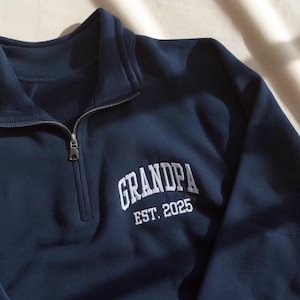 May include: A navy blue quarter-zip sweatshirt featuring white embroidered text on the chest that reads GRANDPA EST. 2025. The left sleeve includes white embroidered text reading EMMA LUKE above a small red heart symbol.