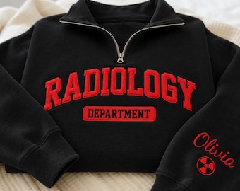 May include: A black quarter-zip sweatshirt featuring the red text RADIOLOGY DEPARTMENT on the chest. The left sleeve displays the name Olivia in red script above a red radiation symbol. The garment has long sleeves with ribbed cuffs and a collar.