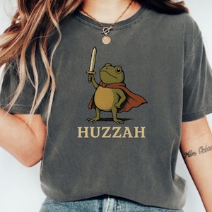 May include: Dark gray t-shirt featuring a cartoon frog with a sword and cape. The frog is holding a sword and wearing a red cape. The word "Huzzah" is printed in gold below the frog.