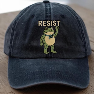 May include: A navy blue baseball cap with the word "RESIST" in tan letters above a cartoon frog illustration. The frog is green with a tan belly and a blue neckerchief, raising a fist.