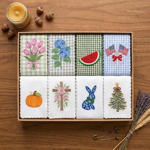May include: A set of eight rectangular fabric towels arranged in a box, featuring embroidered designs including pink tulips, blue hydrangeas, a watermelon slice, two American flags, a pumpkin, a floral cross, a blue rabbit, and a Christmas tree.