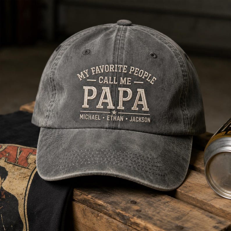 May include: A gray baseball cap with the text "MY FAVORITE PEOPLE CALL ME PAPA" in white embroidery. Below the word "PAPA" are the names Michael, Ethan, and Jackson. The cap is sitting on a wooden surface.