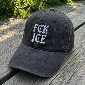 May include: A black baseball cap with the text "FCK ICE" embroidered in white. The cap has a curved brim and a fabric strap closure on the back. The hat is made of a worn, faded material.