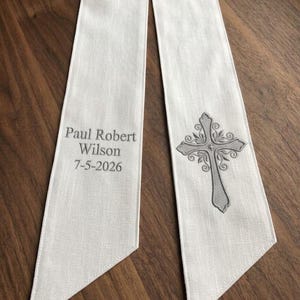 May include: White linen stole with a decorative gray cross and floral design. The stole has the text "Paul Robert Wilson 7-5-2026" printed in gray. The stole is displayed on a wooden surface.