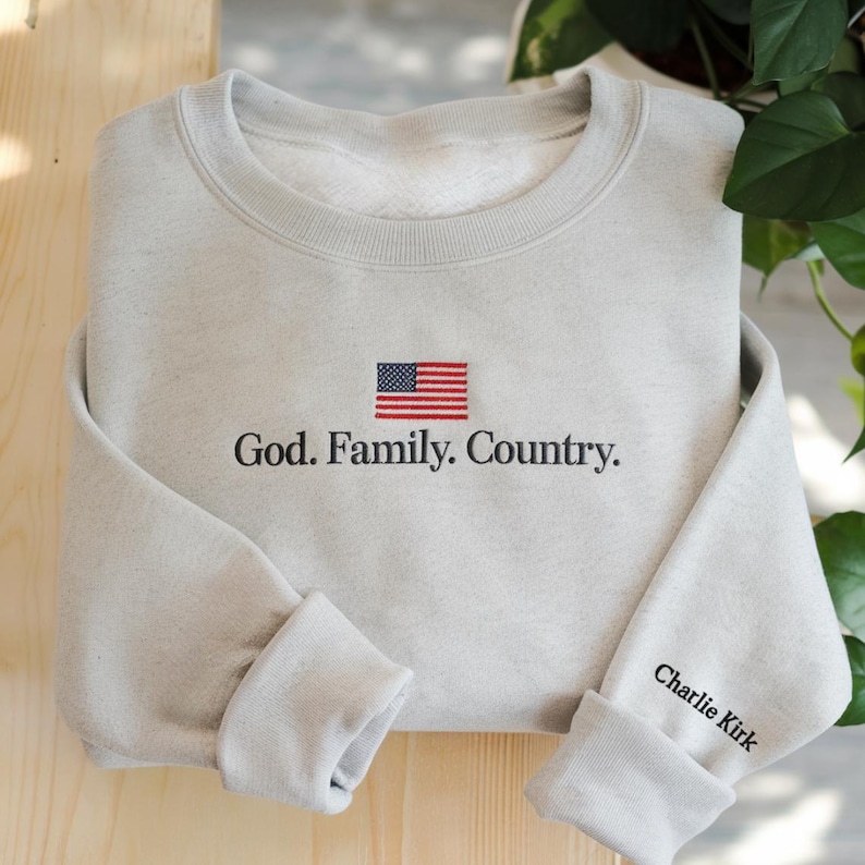 Op de afbeelding: Een lichtgrijs sweatshirt met een geborduurde Amerikaanse vlag en de woorden "God. Family. Country." De rechtermanchet toont de naam "Charlie Kirk." Het sweatshirt is gemaakt van een zacht materiaal. De afbeelding staat op een houten achtergrond.