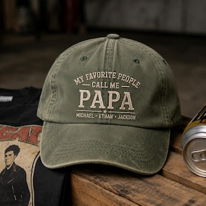 May include: Olive green baseball cap with the text "MY FAVORITE PEOPLE CALL ME PAPA" in white. Below the word "PAPA" are the names "MICHAEL, ETHAN, JACKSON". The hat is on a wooden surface.