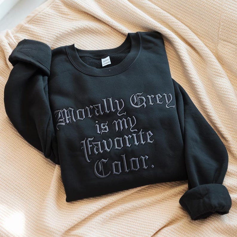 May include: Black sweatshirt with the embroidered text "Morally Grey is my Favorite Color." The long-sleeved top is folded on a beige blanket. The text is in a gothic font.