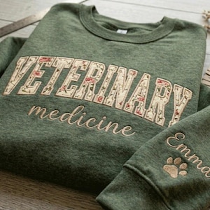 May include: A heathered olive green crewneck sweatshirt featuring the word VETERINARY in large block letters filled with a floral pattern, the word medicine in cursive script, and the name Emma with a paw print embroidered on the sleeve.
