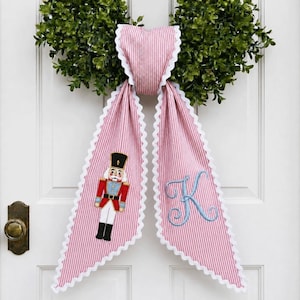 May include: A pink and white pinstriped fabric door hanger featuring a scalloped white trim. One side displays an embroidered nutcracker in a red uniform, while the other side features a large, light blue embroidered letter K in script font.