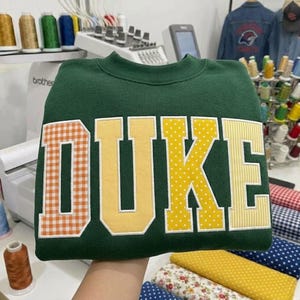 May include: A dark green crewneck sweatshirt featuring the word DUKE in large block letters. Each letter has a unique pattern: the D is orange gingham, the U is solid yellow, the K is yellow with white polka dots, and the E is yellow stripes.