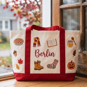 May include: A cream-colored canvas tote bag with red handles and a red base. The bag features embroidered autumn-themed icons including a pie, scarf, open book, honey jar, acorn, leaves, boots, striped sock, and a jack-o'-lantern, with the name Berlin.