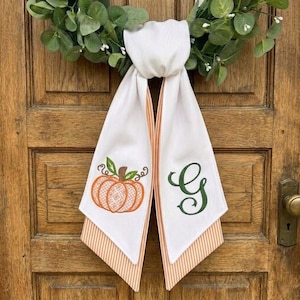 May include: A white fabric door hanger featuring a green eucalyptus wreath at the top. The two hanging tails display an embroidered orange pumpkin with green leaves on the left and a large, dark green cursive letter G on the right side.