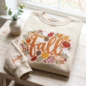 May include: A beige crewneck sweatshirt featuring an embroidered design of the word fall in cursive script, surrounded by pumpkins, pinecones, autumn leaves, and berries. The name Jenifer is embroidered in orange thread on the left sleeve cuff.
