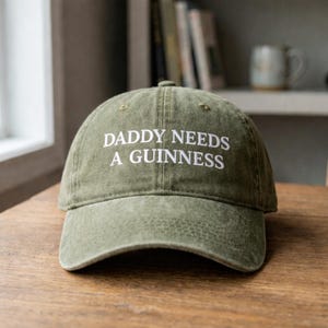 May include: Olive green baseball cap with the embroidered text "DADDY NEEDS A GUINNESS" in white. The cap is sitting on a wooden surface. The background includes a window, books, and a mug.