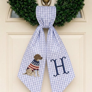 May include: A light blue and white gingham fabric door hanger featuring an embroidered brown dog wearing a red, white, and blue star-patterned sweater on one side and a large, dark blue embroidered letter H on the other side of the fabric.