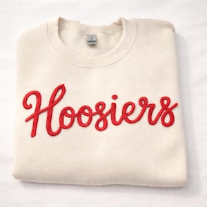 May include: Cream-colored sweatshirt with the word "Hoosiers" embroidered in red script across the front. The sweatshirt is folded neatly, showcasing the design. The garment has a crew neck and a small tag.