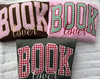 May include: Three sweatshirts featuring the text BOOK in large block letters with a gingham pattern and the word lover in cursive script. One is brown with pink gingham, one is pink with green gingham, and one is dark gray with red gingham.