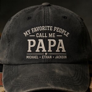 May include: A dark gray baseball cap with the embroidered text "MY FAVORITE PEOPLE CALL ME PAPA" in white. Below, the names MICHAEL, ETHAN, and JACKSON are also embroidered. The cap has a curved brim and a button on top.