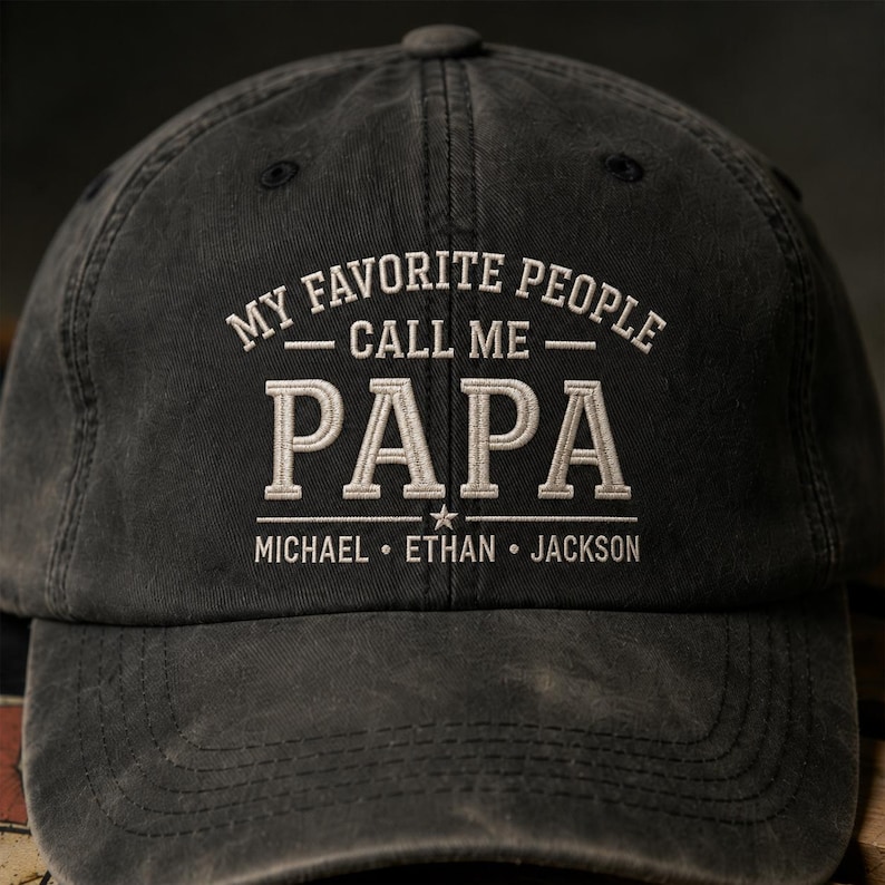 May include: A dark gray baseball cap with the embroidered text "MY FAVORITE PEOPLE CALL ME PAPA" in white. Below, the names MICHAEL, ETHAN, and JACKSON are also embroidered. The cap has a curved brim and a button on top.