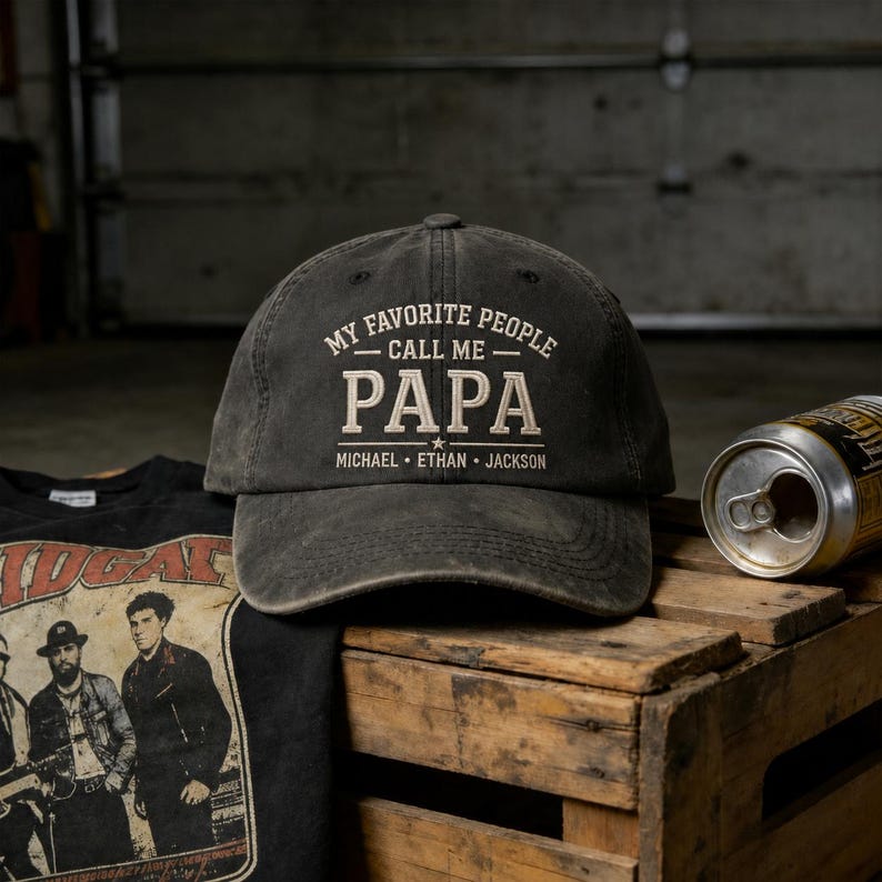 May include: A dark gray baseball cap with the text "MY FAVORITE PEOPLE CALL ME PAPA" in white. Below the word "PAPA" are the names "MICHAEL ETHAN JACKSON". The cap is sitting on a wooden crate next to a black t-shirt and a can.