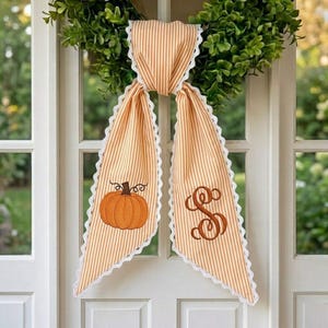 May include: A decorative door hanger featuring a green leaf wreath adorned with two long, light orange and white striped fabric tails. The left tail displays an embroidered orange pumpkin, while the right tail features a brown embroidered letter S.
